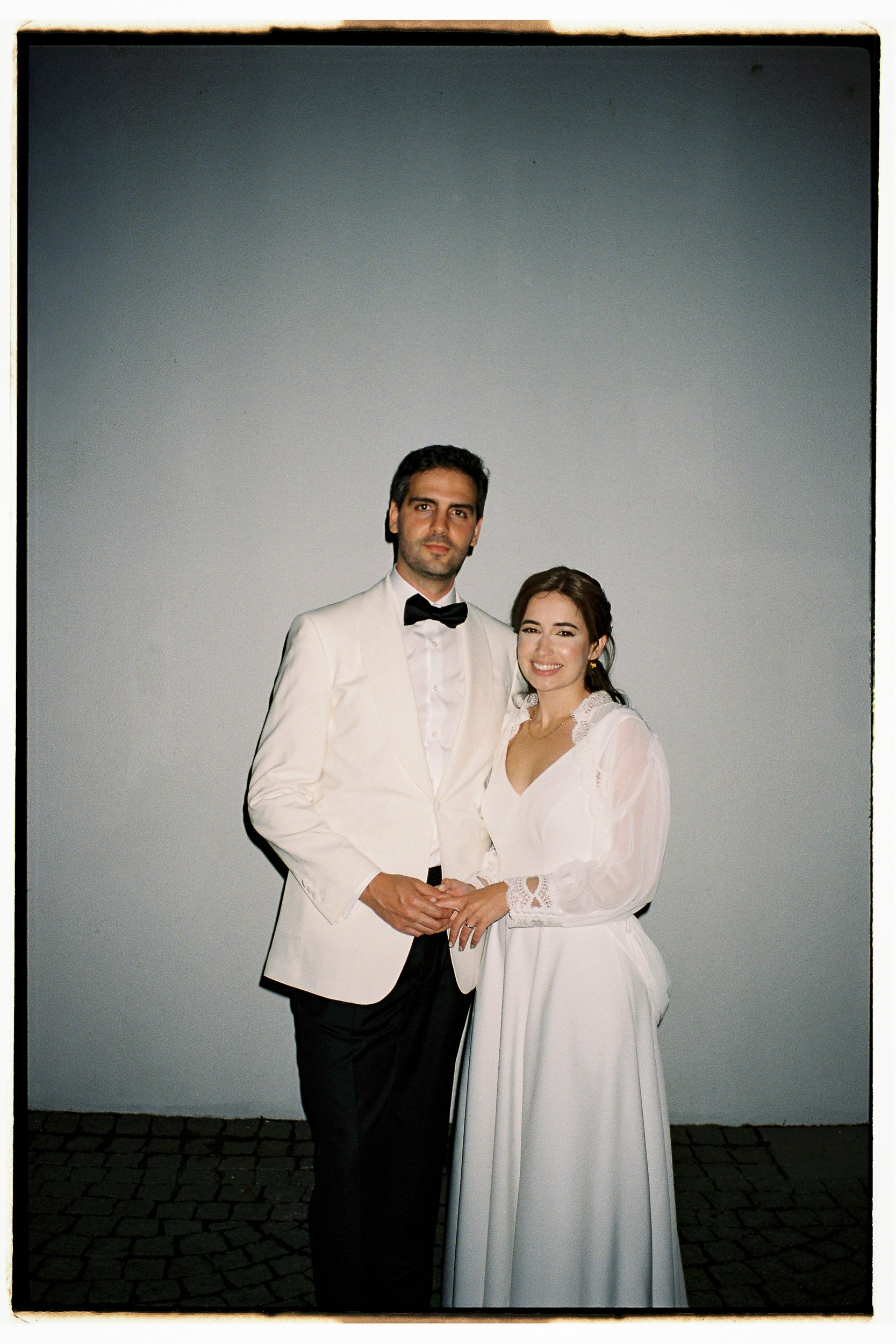 A couple in wedding attire standing outdoors on a cobblestone street against a plain wall. The man is wearing a white tuxedo with a black bow tie, and the woman is in a white wedding dress with lace details and a veil. They are holding hands and smiling at the camera.