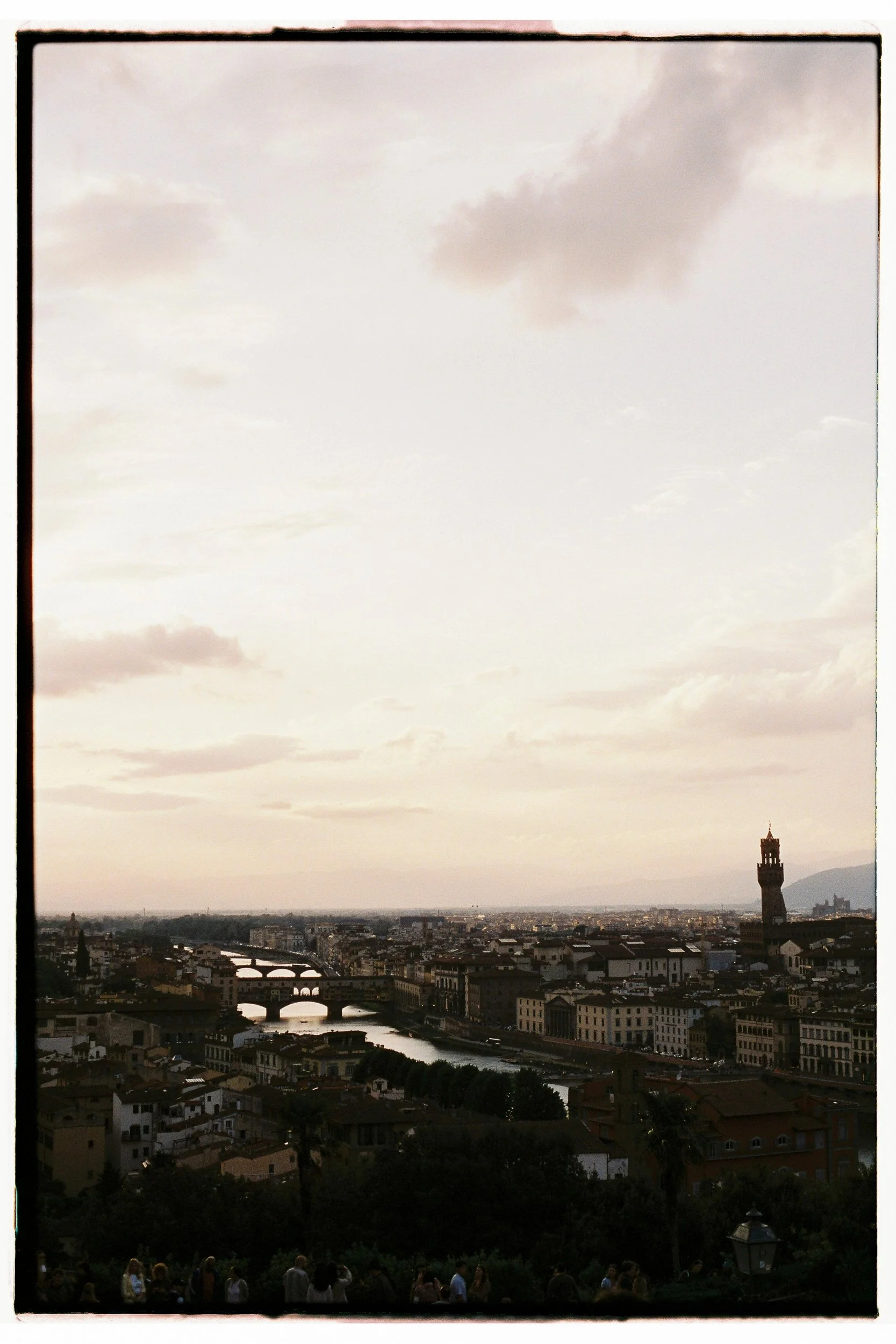 A city skyline during sunset with a river running through it, and a tall tower on the right side. There are clouds in the sky and people are gathered in a park at the foreground.