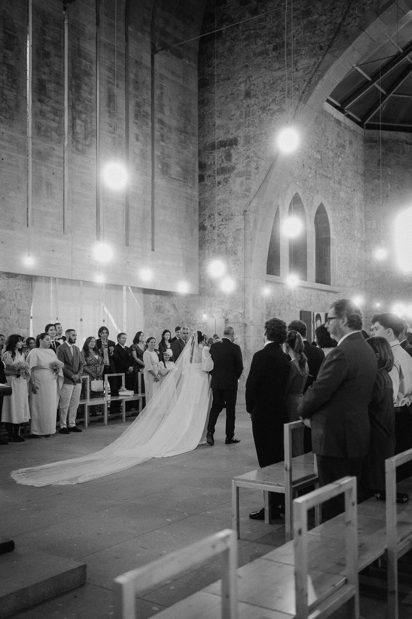 Black and white photograph of a wedding ceremony in a church, with the bride and groom standing at the altar, surrounded by guests seated on wooden benches, with high arched windows and stone walls.