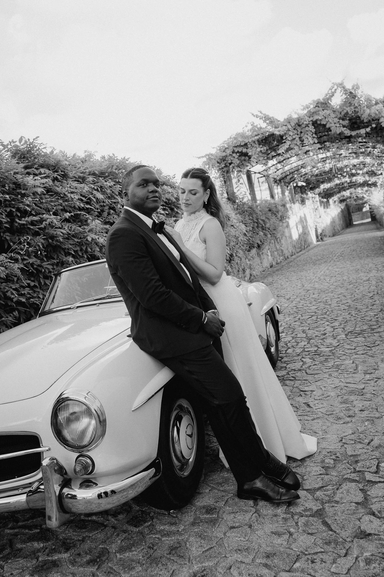 A black and white photo of a man and woman dressed in formal wedding attire, posing beside a vintage white car on a cobblestone street with a lush, leafy backdrop and an arched trellis overhead.