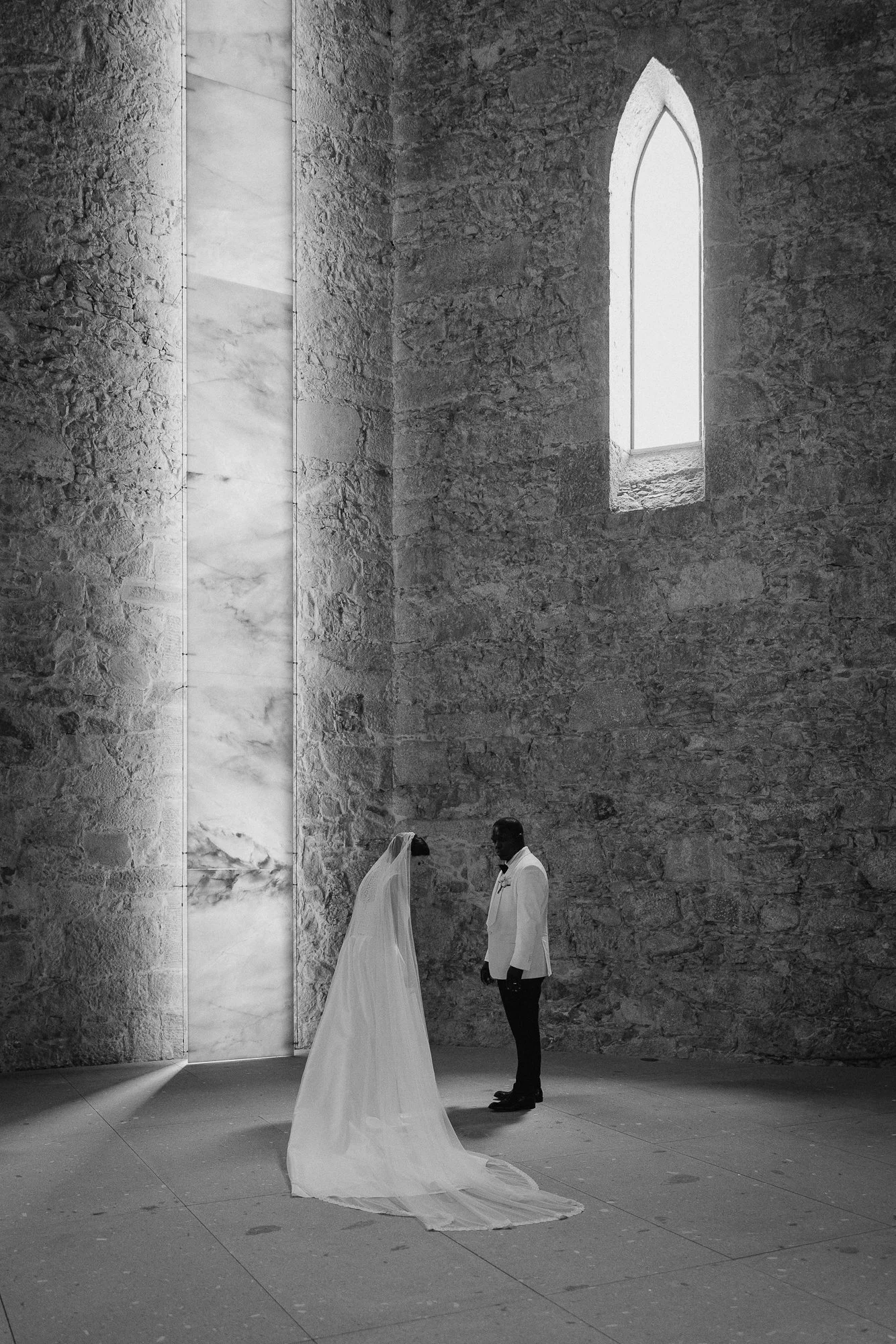 A bride in a wedding gown and a groom in a tuxedo standing inside a stone church with high arched windows and textured walls.