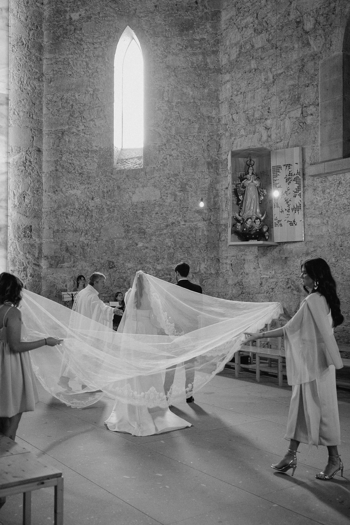 People participating in a wedding ceremony inside a stone church with a tall arched window and religious statue on the wall.