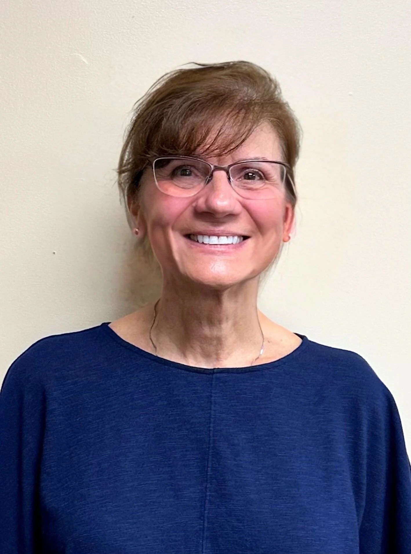 A smiling woman with short brown hair, glasses, and a blue top standing against a plain light-colored wall.