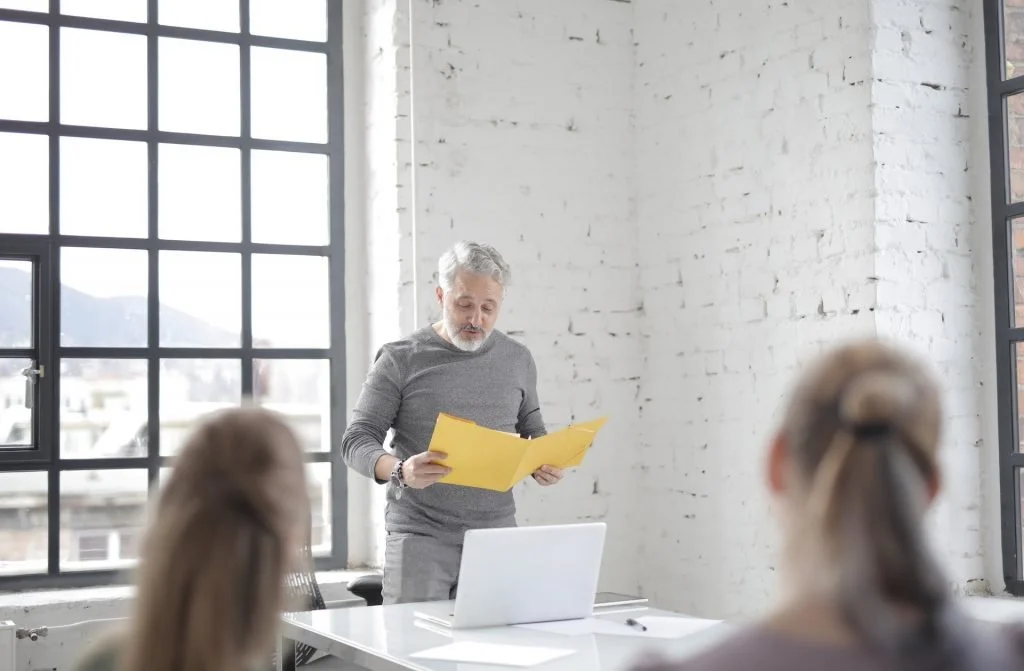 corporate-man-presenting-yellow-folder-two-women-collegues-office-window-white-brick-wall