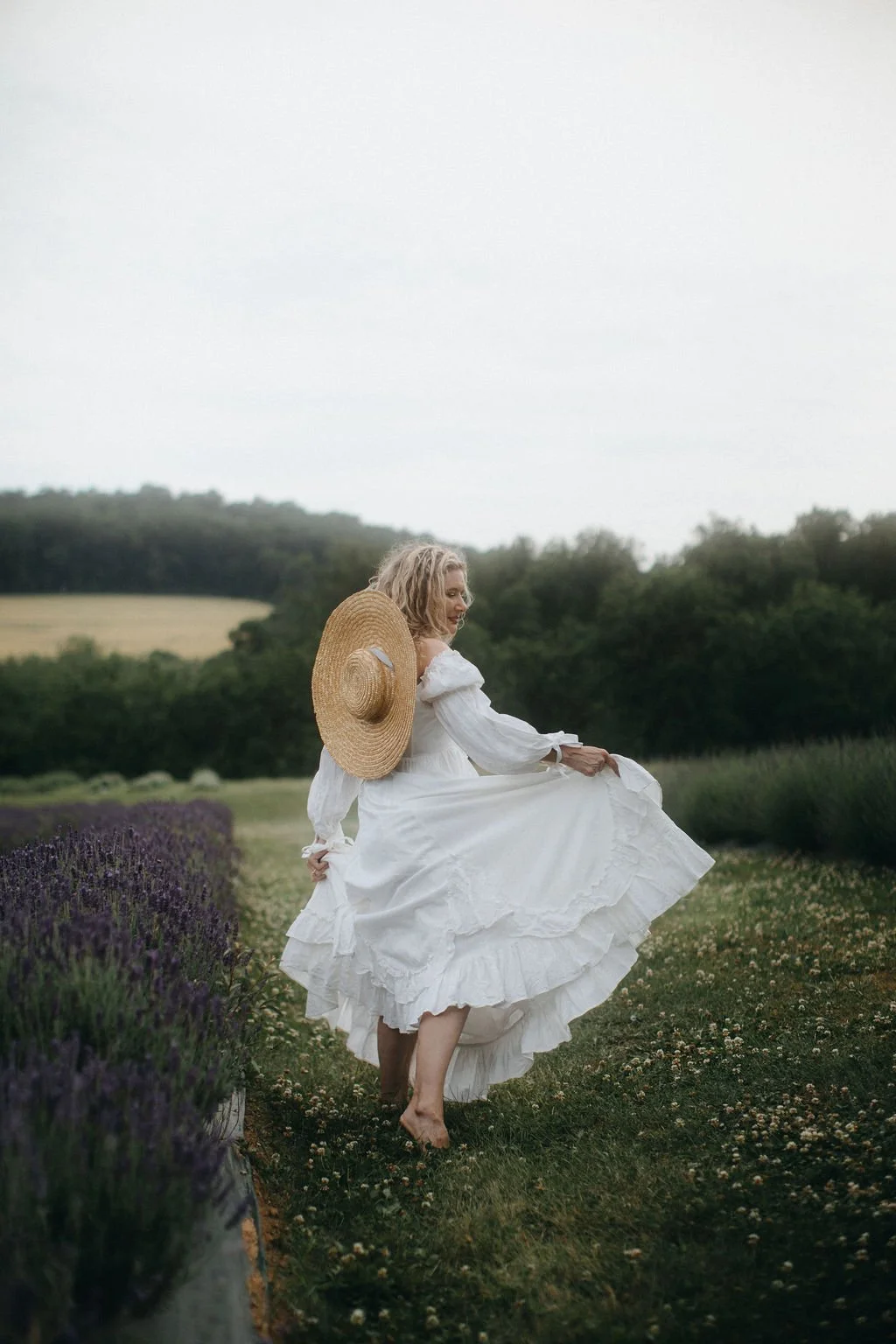 Kim, a shamanic practitioner, walks barefoot looking over her shoulder. She is wearing a striking, vintage, long white dress in a garden full of lavender and greenery.