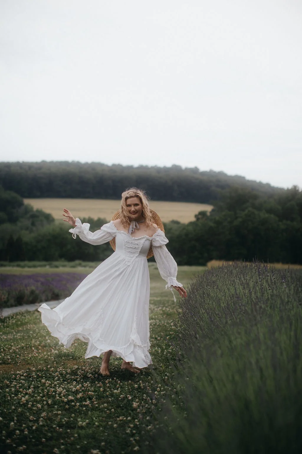 Kim, a shamanic practitioner, faces the camera in a white vintage dress standing in a field of lavender, with rolling green hills in the background, smiling.