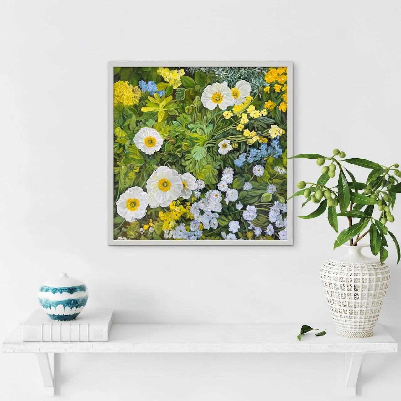 Print of white, blue and yellow flowers in green garden, framed in white, on white wall above white shelf with book, vase and green leaves