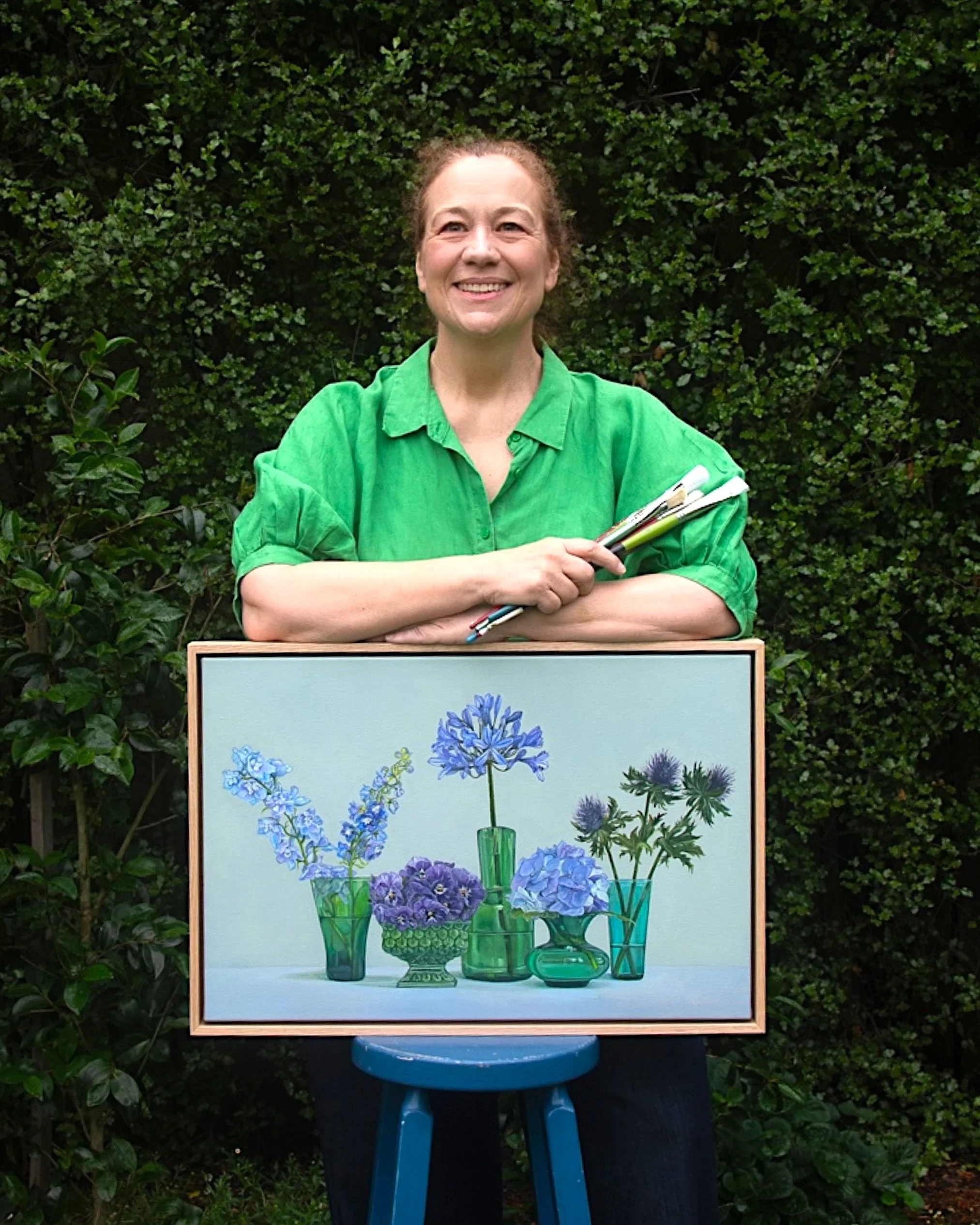 Robyn Markey in green shirt holding paint brushes and leaning on oil painting of green vases filled with flowers