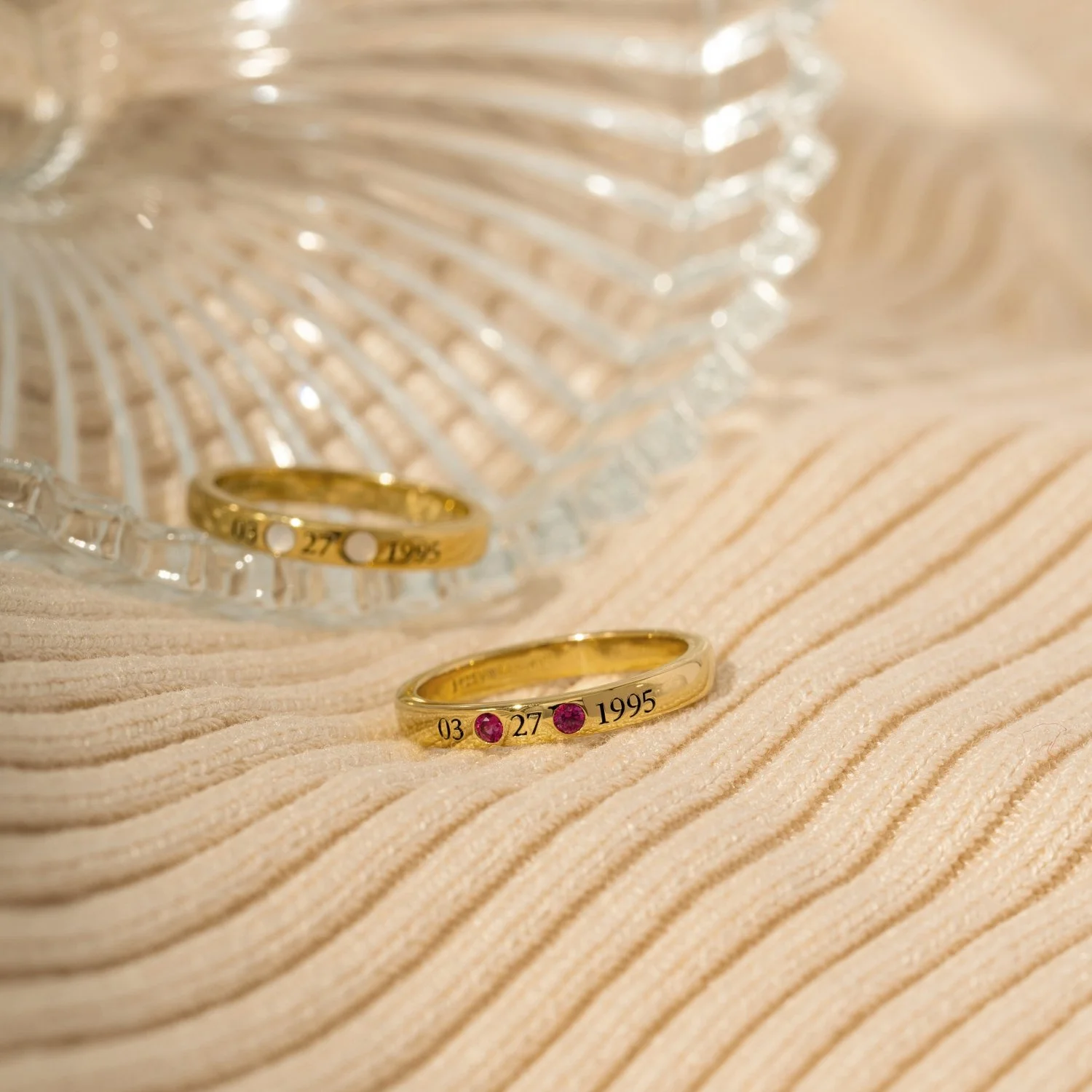 Close-up of two gold rings engraved with date and numbers, placed on a textured beige surface, with a glass bowl nearby.