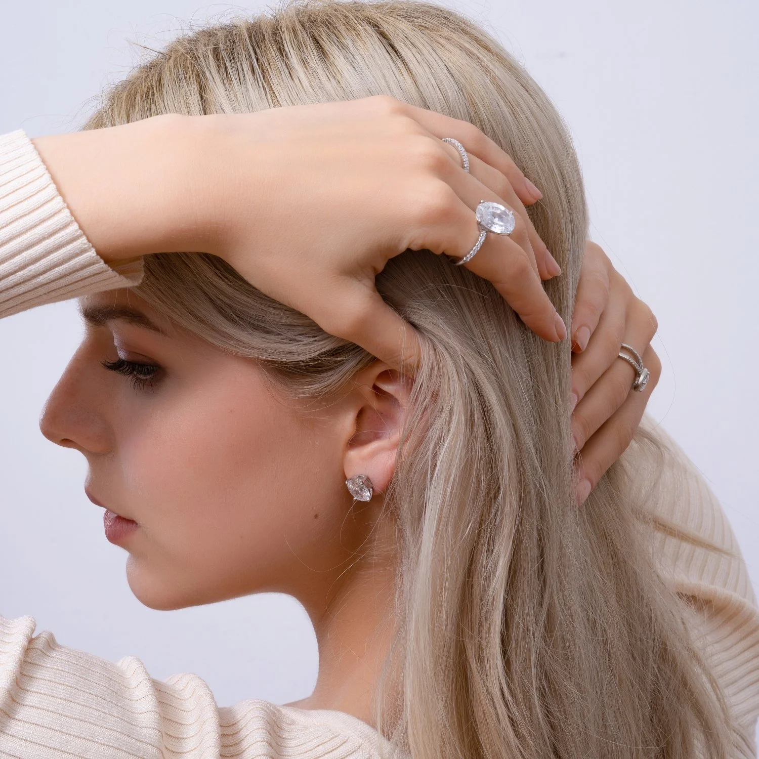 A woman with blonde hair and diamond jewelry, including earrings and rings, in a side profile pose with one hand on her head and the other behind her ear.