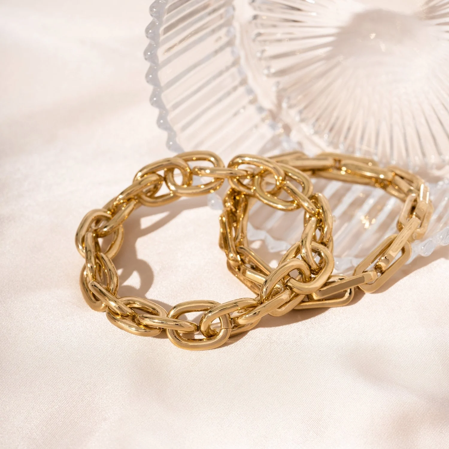 Three gold chain bracelets and a gold chain ring on a white surface with a glass dish in the background.