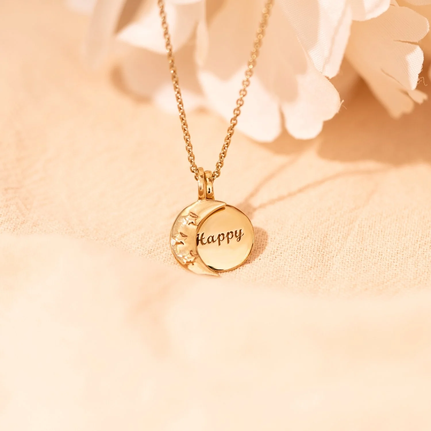 Gold necklace with a round pendant engraved with the word 'Happy' and a crescent moon design, resting on a soft pink surface near white flowers.