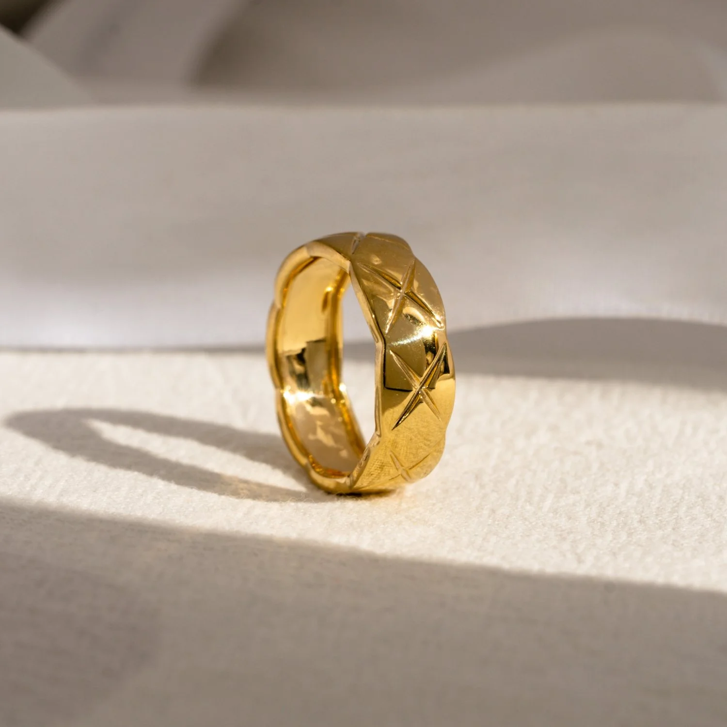 Close-up of a gold ring with a textured surface and engraved star patterns, placed on a white textured surface with a shadow cast behind it.