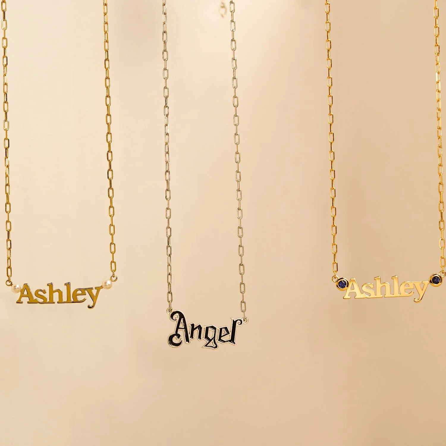 Three necklaces with names: Ashley and Angel, on a beige background. The left and right necklaces have names in gold with small pearls or gemstones, and the middle necklace has the name Angel in black with a silver chain.