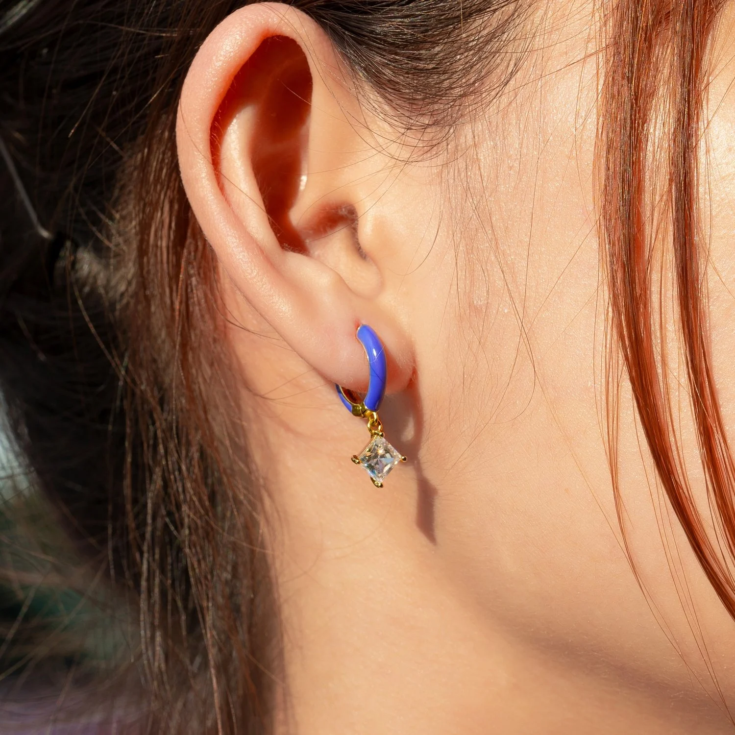 Close-up of a woman's right ear wearing a gold hoop earring with a blue enamel finish and a dangling square-shaped crystal pendant.