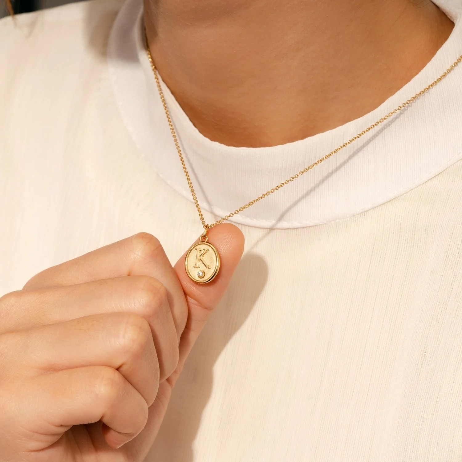 Close-up of a person wearing a white shirt and holding a gold necklace with a pendant engraved with the letter 'K' and a small gemstone.