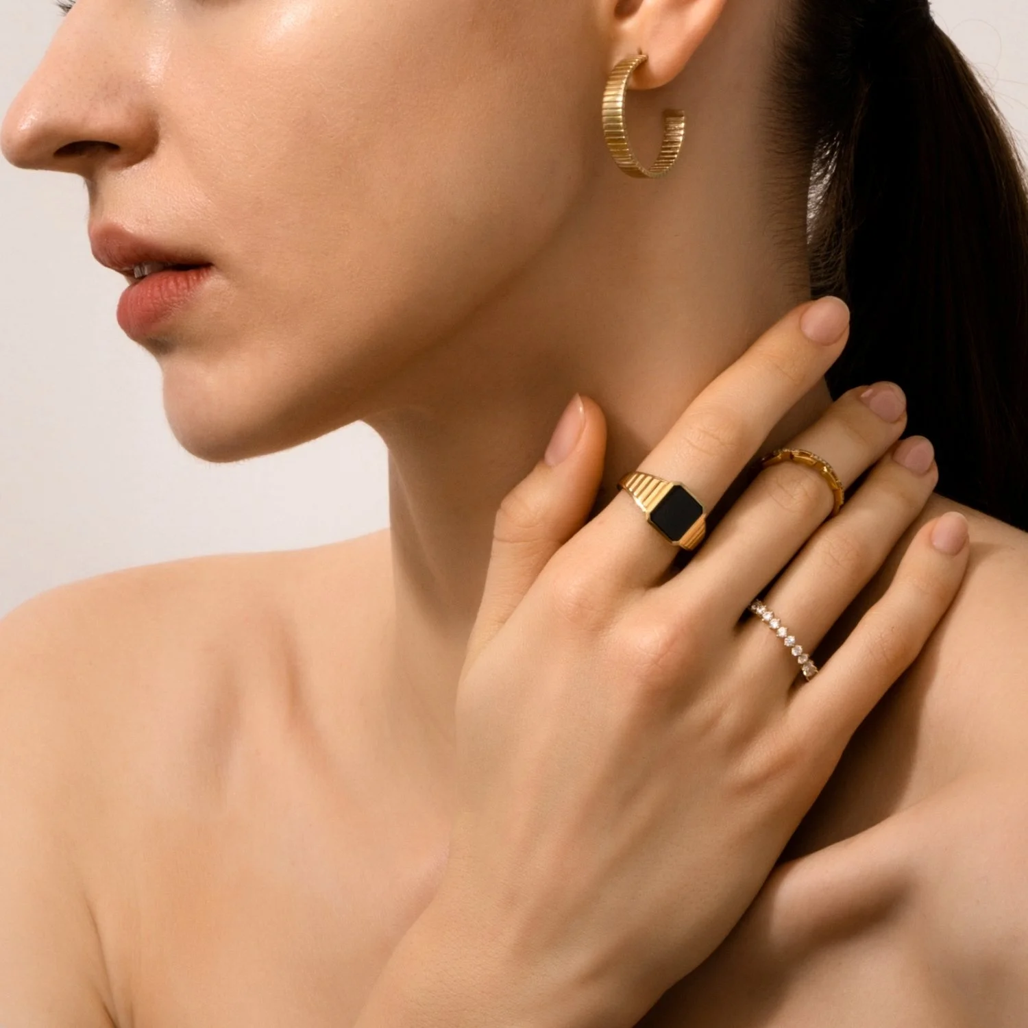 Close-up of a woman's face and hand, showcasing gold jewelry including ring earrings and rings, with a neutral background.
