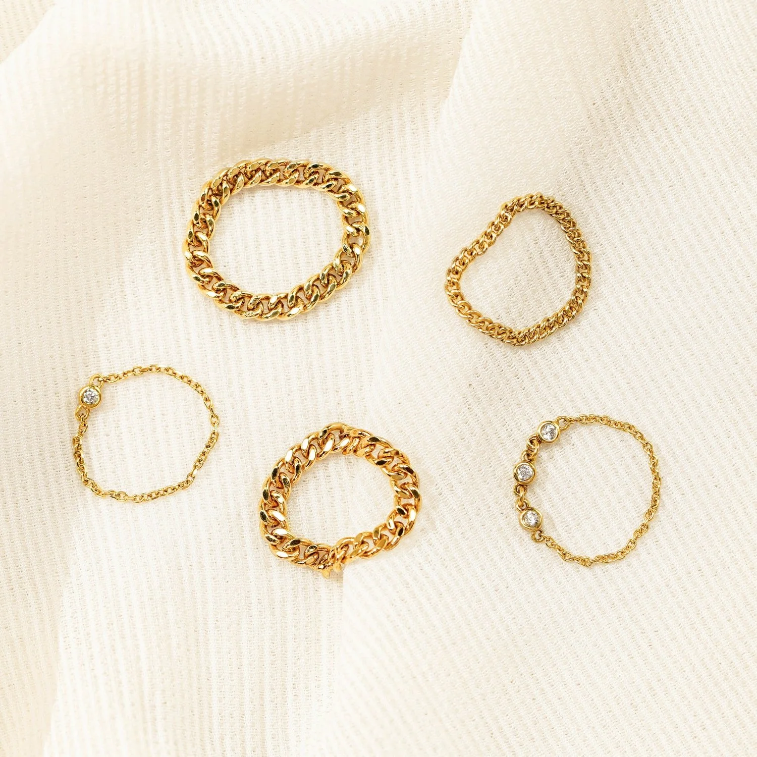 Gold jewelry including two thick chain link rings, one thin chain ring with diamonds, and one slightly textured chain link ring, all arranged on a light beige fabric background.