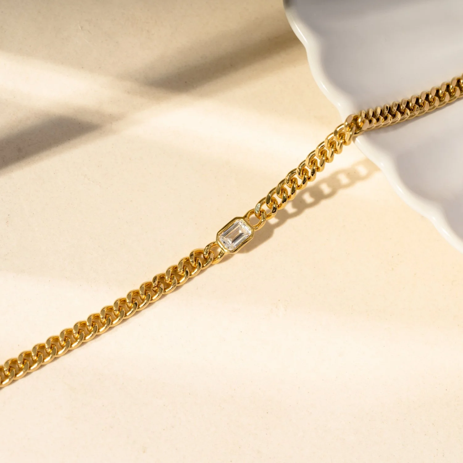Gold chain bracelet with a rectangular clear gemstone, placed on a cream surface near a white ceramic dish.