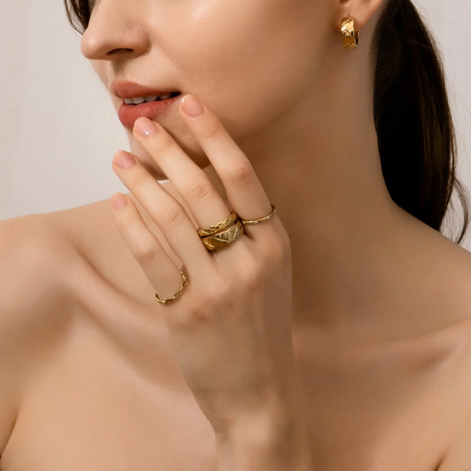 Close-up of a woman with gold jewelry including earrings and rings, touching her lips with fingers, showing bare shoulder and neck against a plain background.