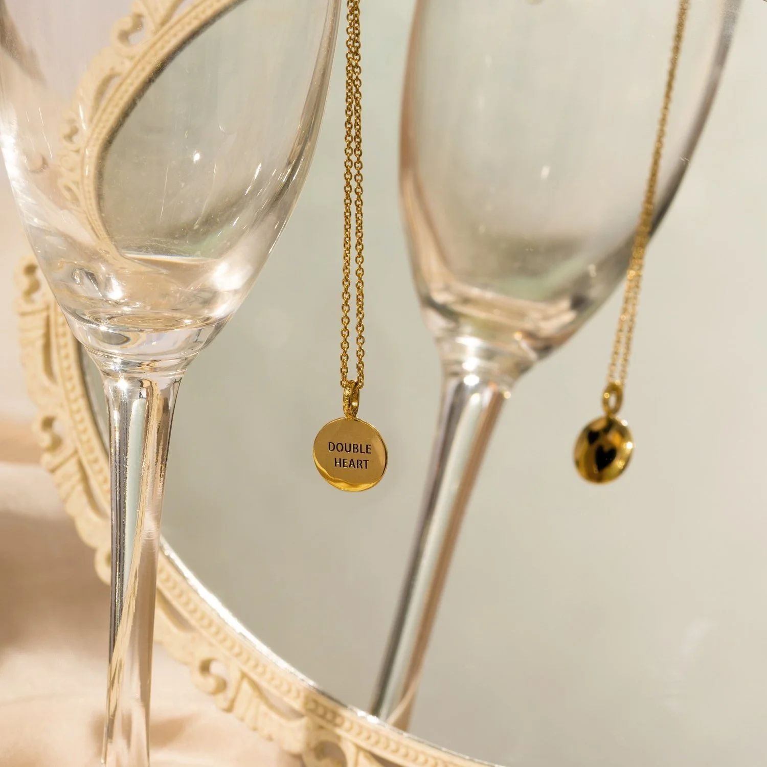 Two champagne flutes with gold necklaces hanging inside, one with a pendant labeled 'DOUBLE HEART' and the other with a small black and gold ladybug charm, reflected in a round mirror with an ornate white frame.
