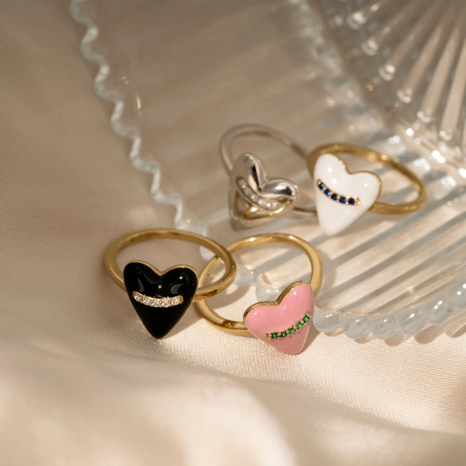 Five rings with heart-shaped designs, two gold, one silver, and two with enamel in black and pink, decorated with small gemstones, displayed on a glass dish.