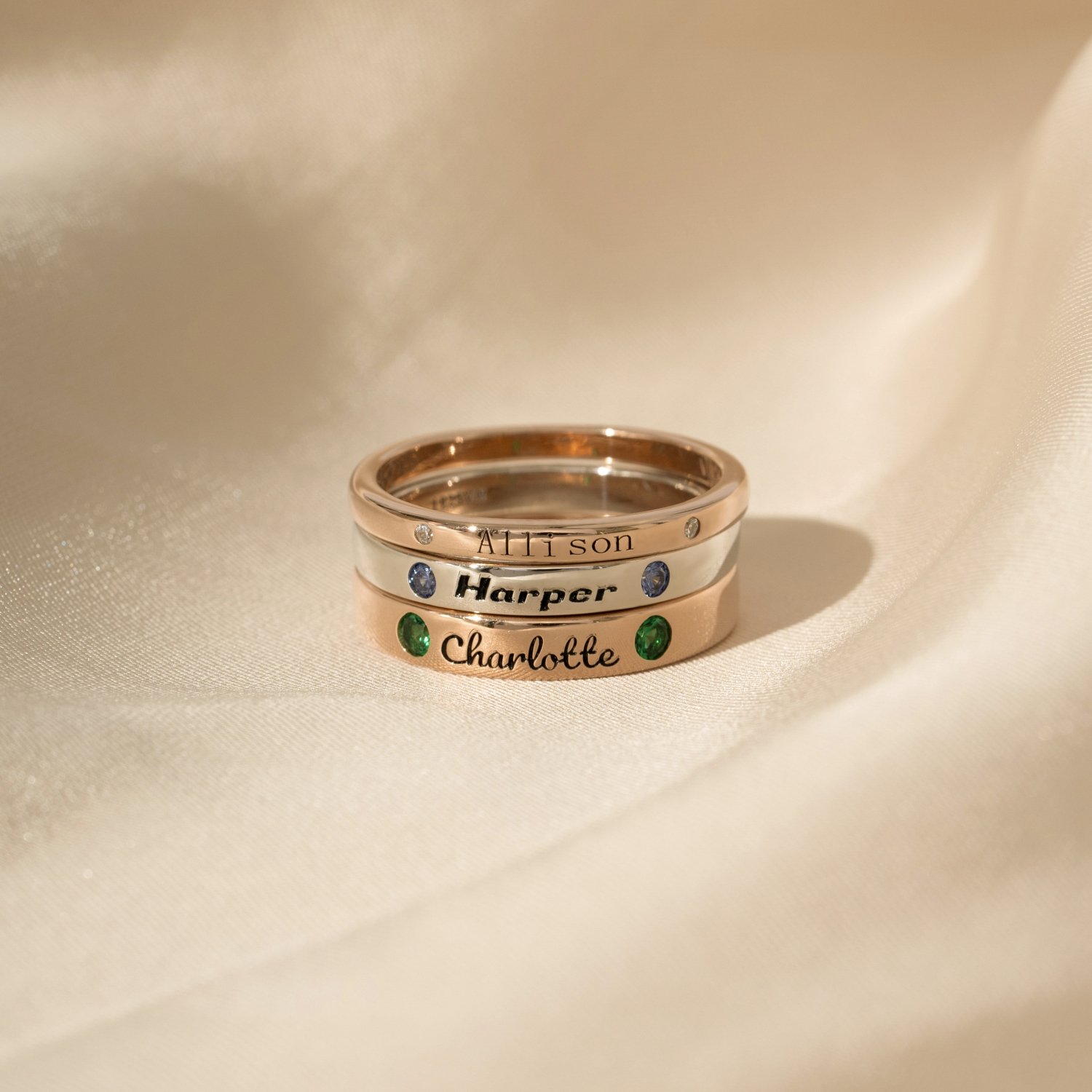 Three rings engraved with the names Allison, Harper, and Charlotte, with gemstones in blue, green, and no color respectively, on a silky beige fabric background.
