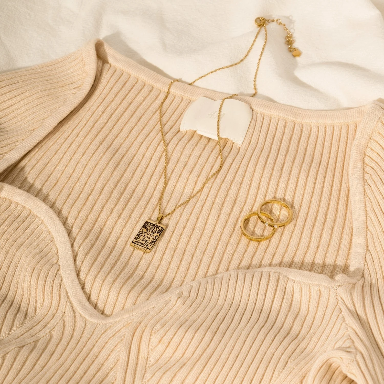 White ribbed sweater with jewelry including a gold chain necklace with a pendant, a bracelet, and two rings, all laid out on a bed or fabric surface.