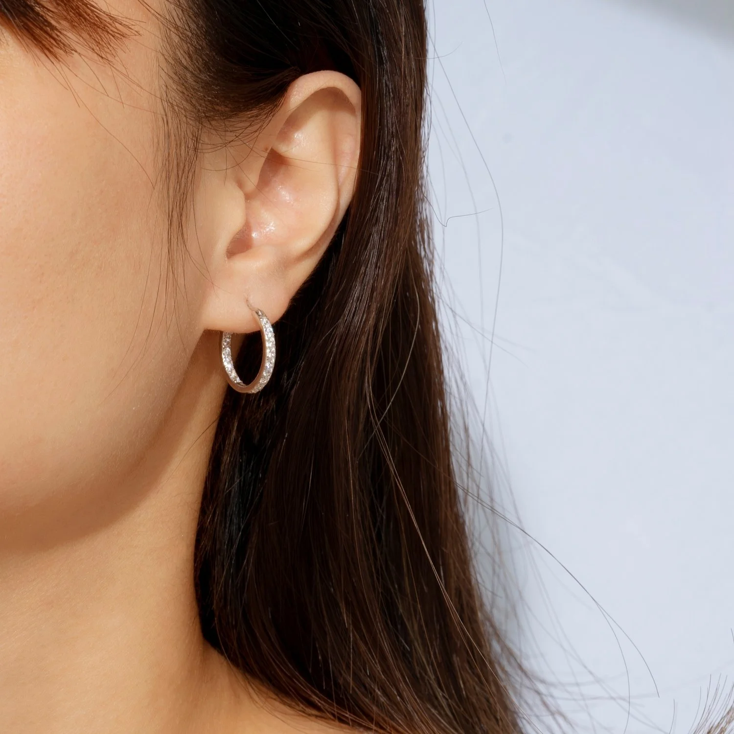 Close-up of a woman's ear wearing a silver hoop earring with small rhinestones, with brown hair partly covering the ear.