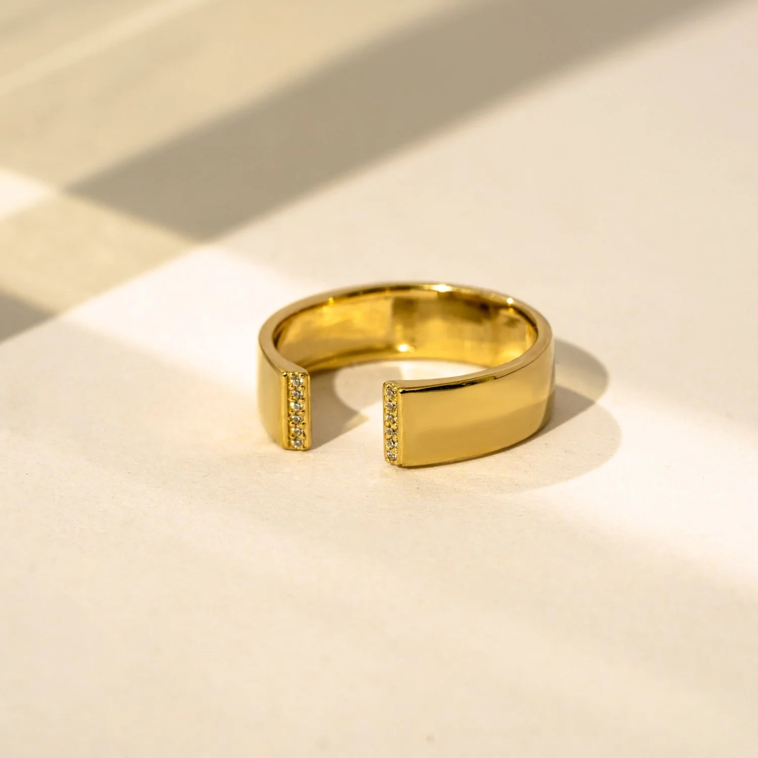 Gold open-ended ring with small embedded diamonds on both ends, placed on a white surface with shadows indicating lighting.