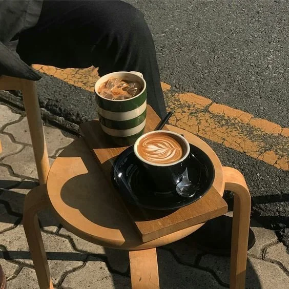 Two cups of coffee, one with a latte art design, on a small wooden table outside on a sidewalk.
