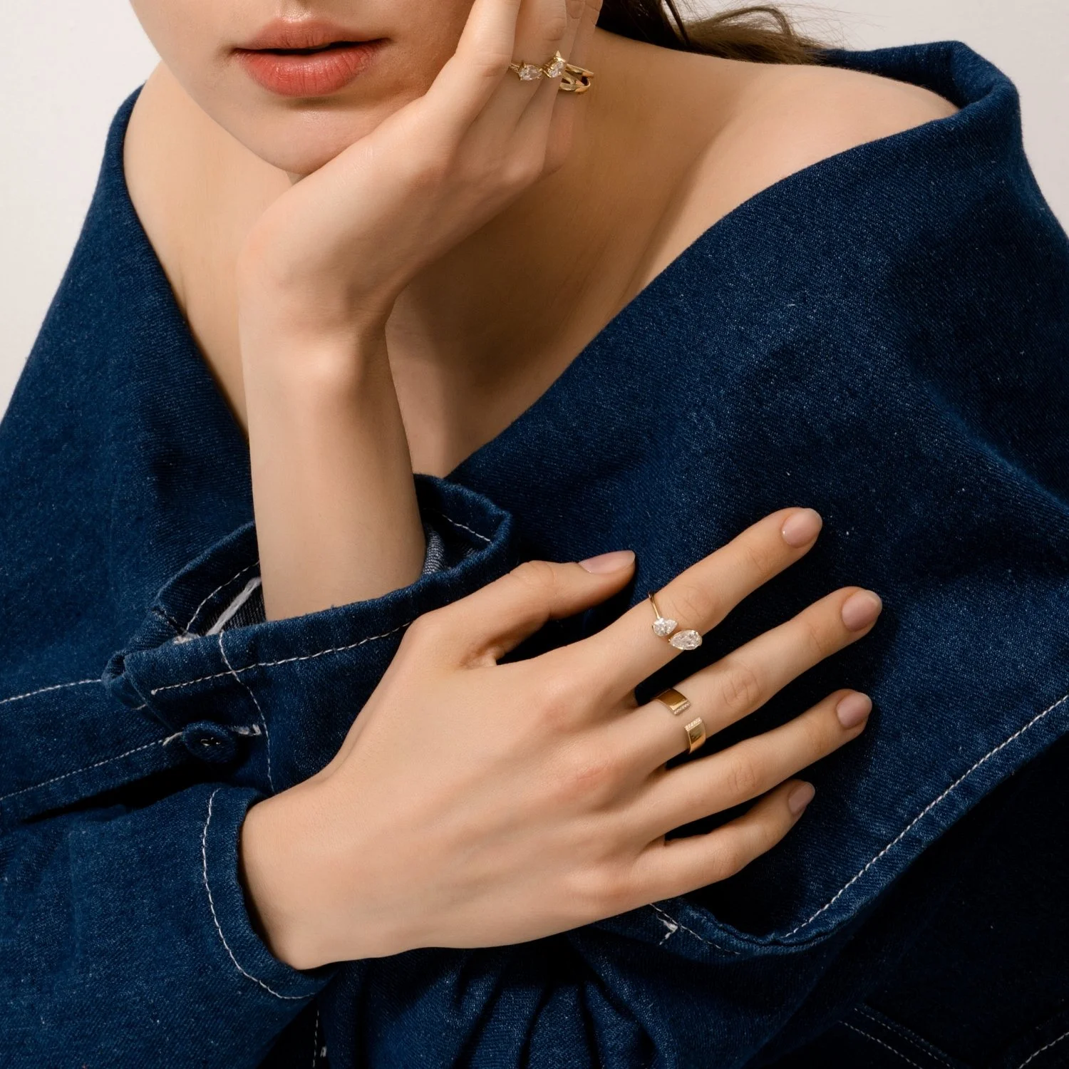 Close-up of a woman wearing denim jacket, showing her hands with jewelry, including a ring with two marquise-cut stones, another ring, and a gold band, while resting her chin on her hand.
