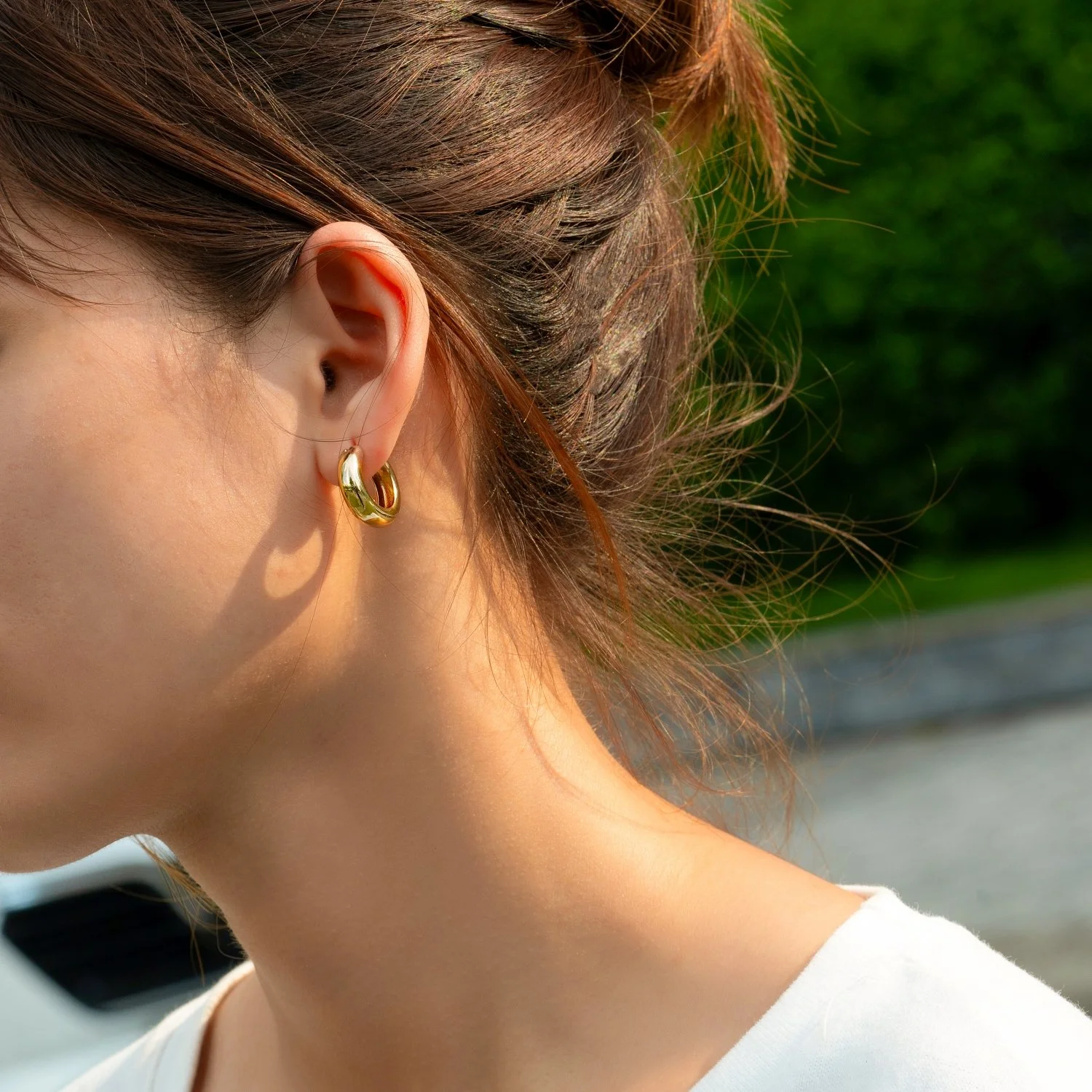 Close-up of a woman's ear with a gold hoop earring, and her brown hair pulled back, outdoors with blurred green background.