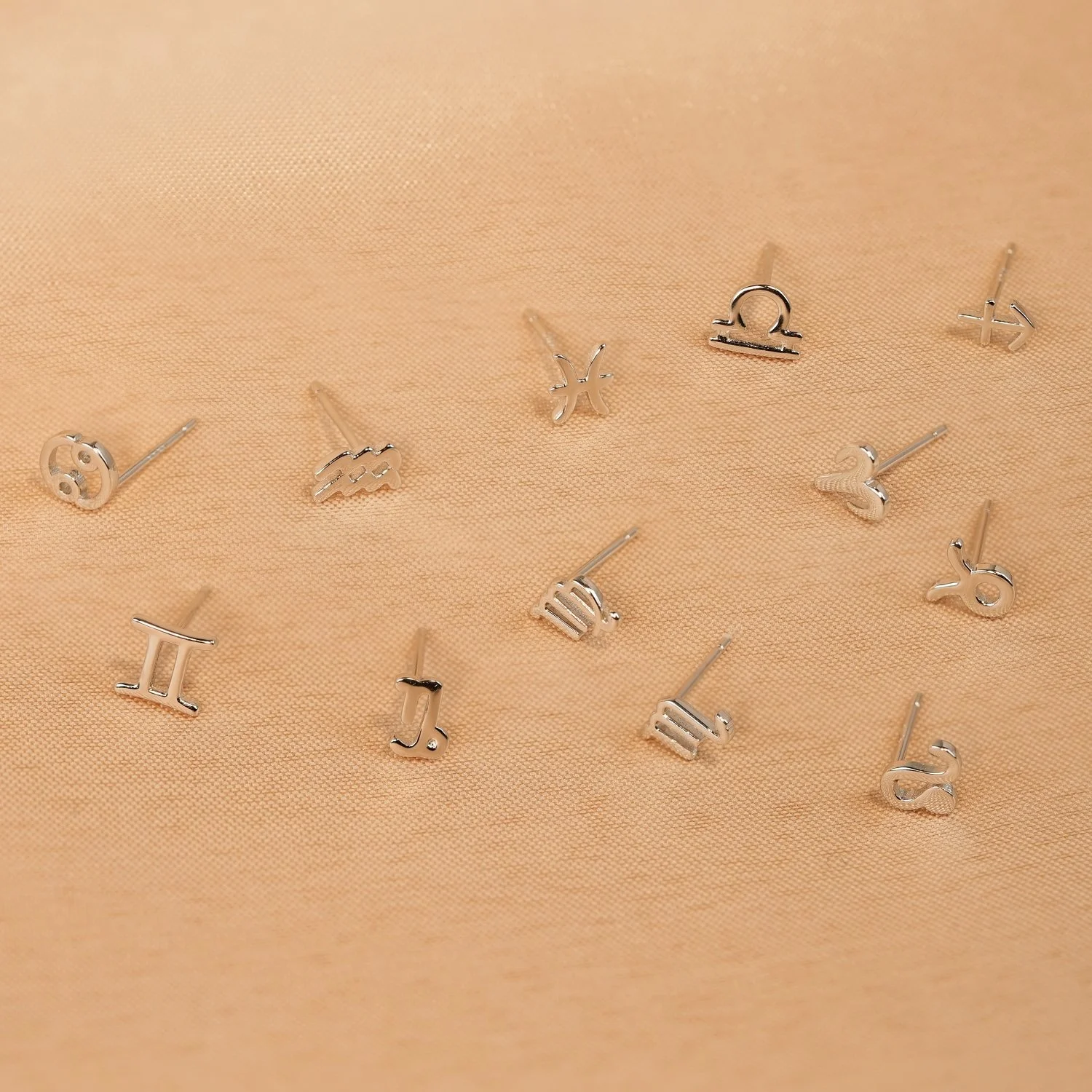 Collection of small silver zodiac sign earrings on peach-colored fabric.