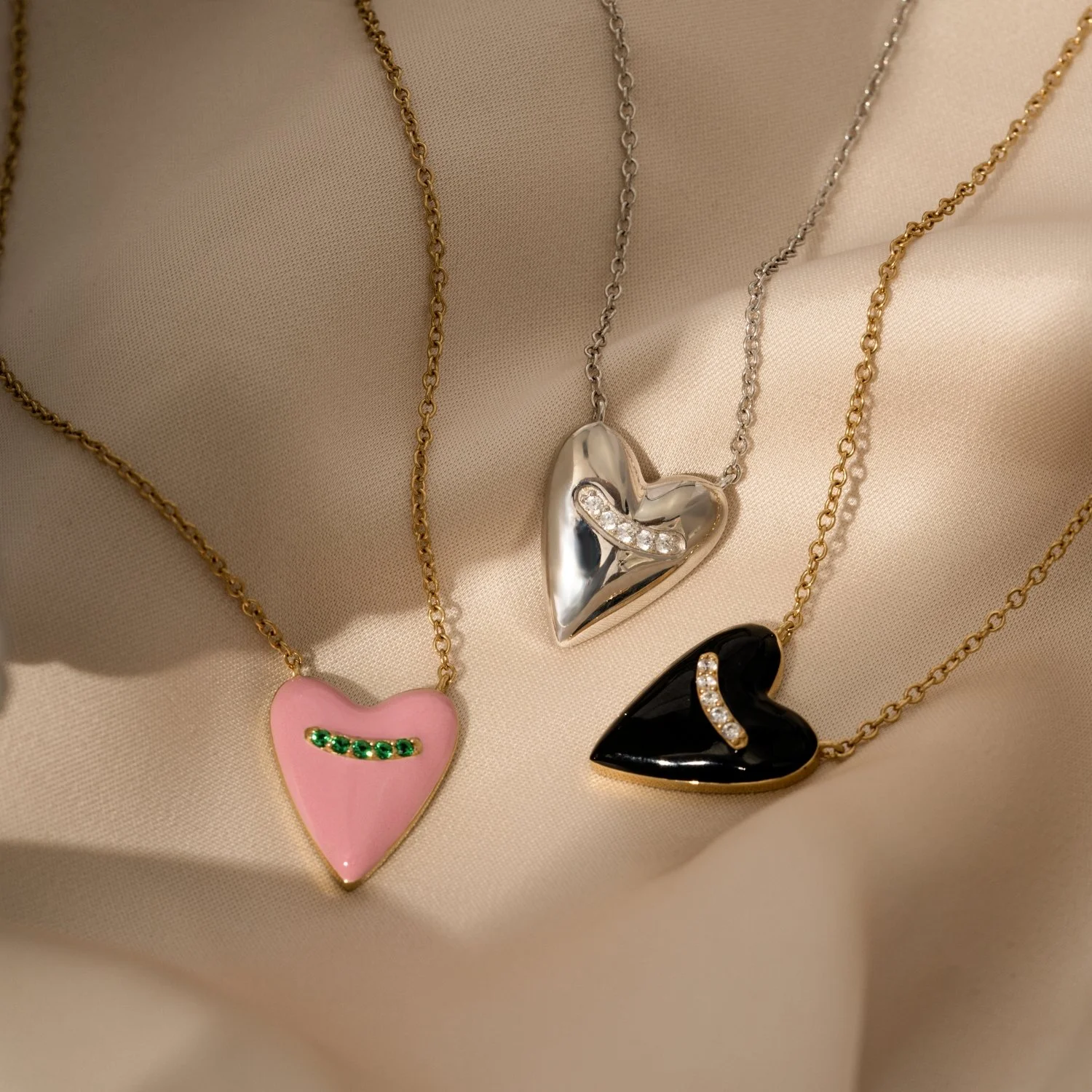 Three necklaces with heart-shaped pendants on a beige fabric background. The left pendant is pink with green gemstones, the middle pendant is silver with a curved row of clear gemstones, and the right pendant is black with a curved row of clear gemstones.