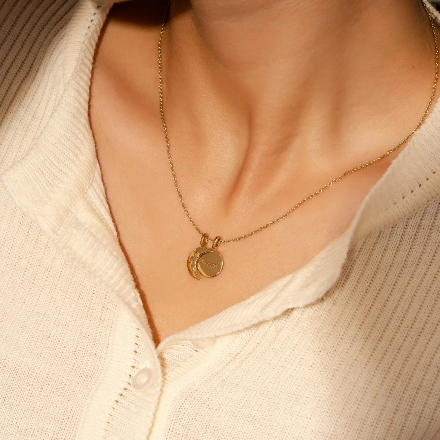 A close-up of a woman wearing a gold necklace with a round pendant that has a moon and star design, and the word 'happy' engraved on it. She is dressed in a cream-colored, textured button-up shirt.