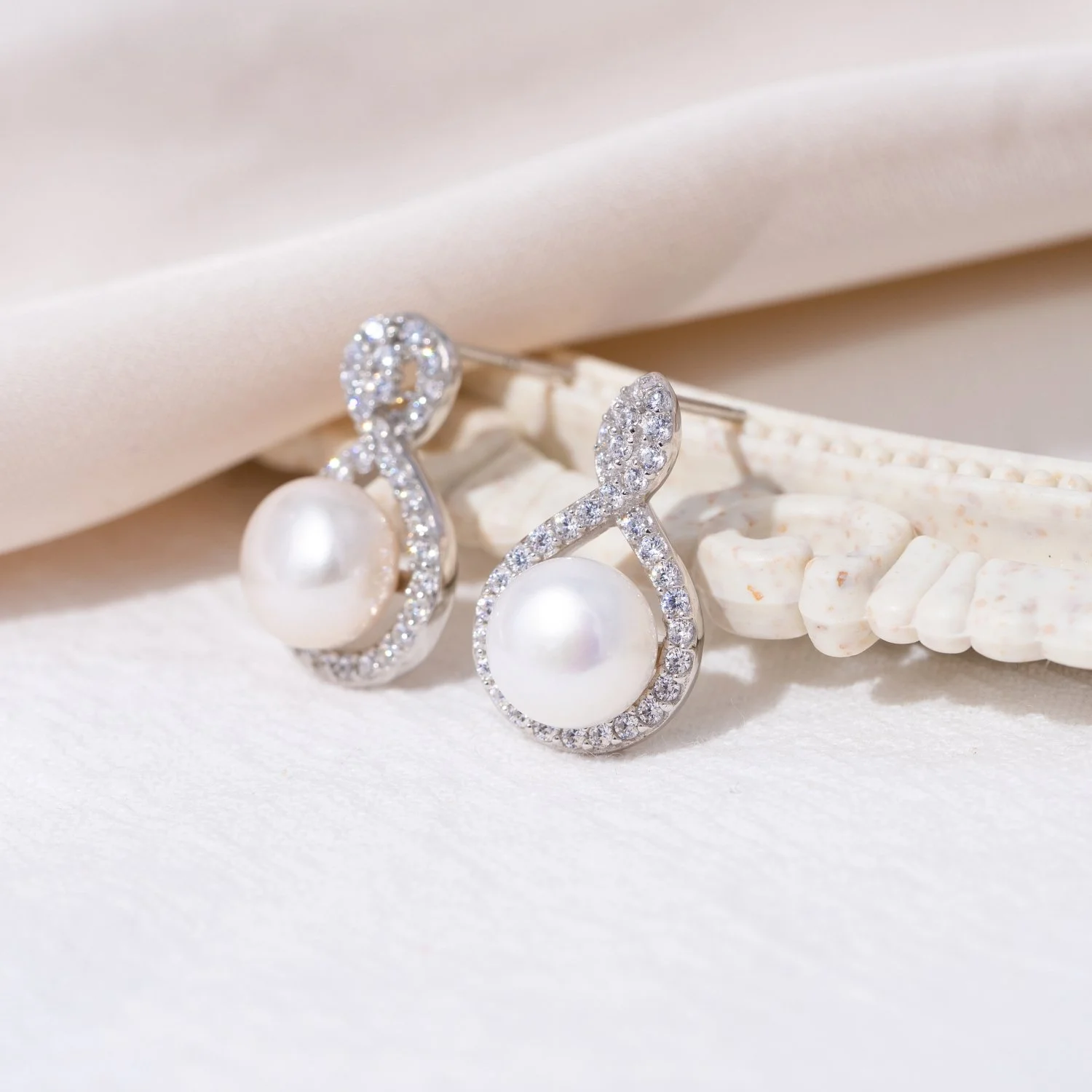 Silver earrings with large white pearls and encrusted with small diamonds, placed on a soft white surface with a piece of white fabric and seashell in the background.