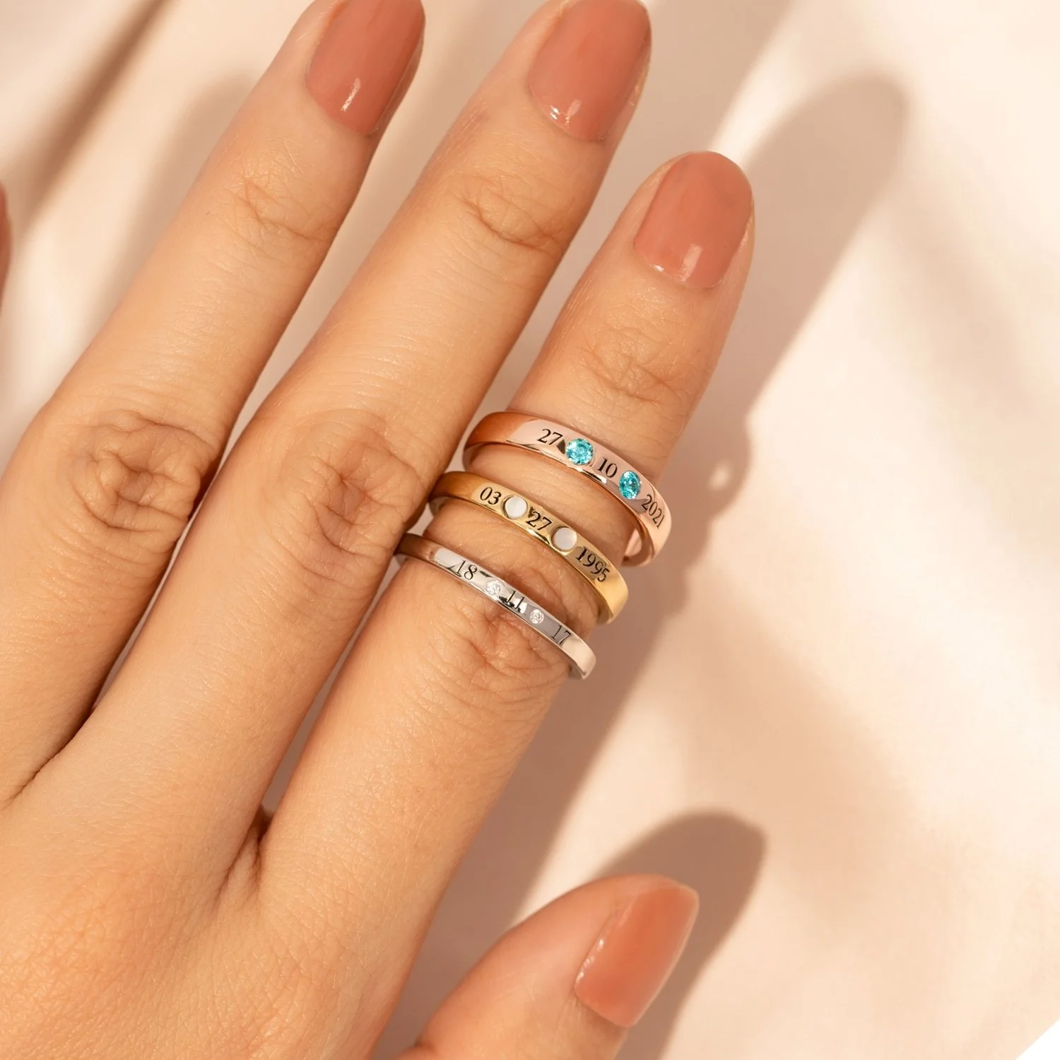 A hand with three stacked rings on the index finger. The rings have small attached inscriptions with dates and symbols. The background is a soft, creamy fabric.