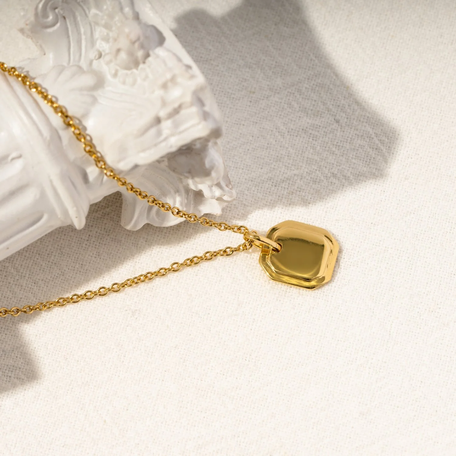 Gold necklace with a hexagonal pendant resting on a white decorative object on a light-colored surface.