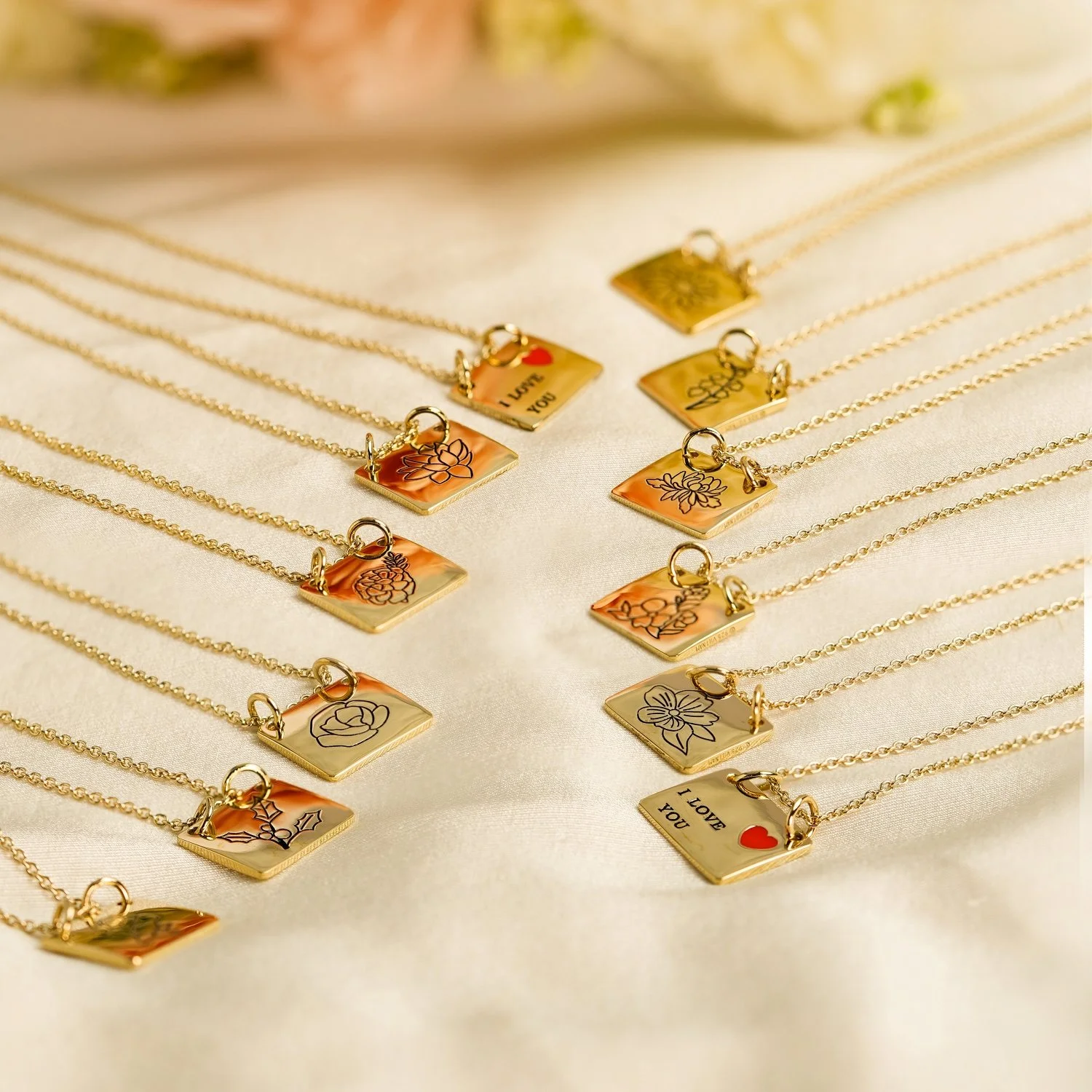 Gold necklaces with square pendants featuring various engraved floral and love-themed designs, laid on a light fabric surface.