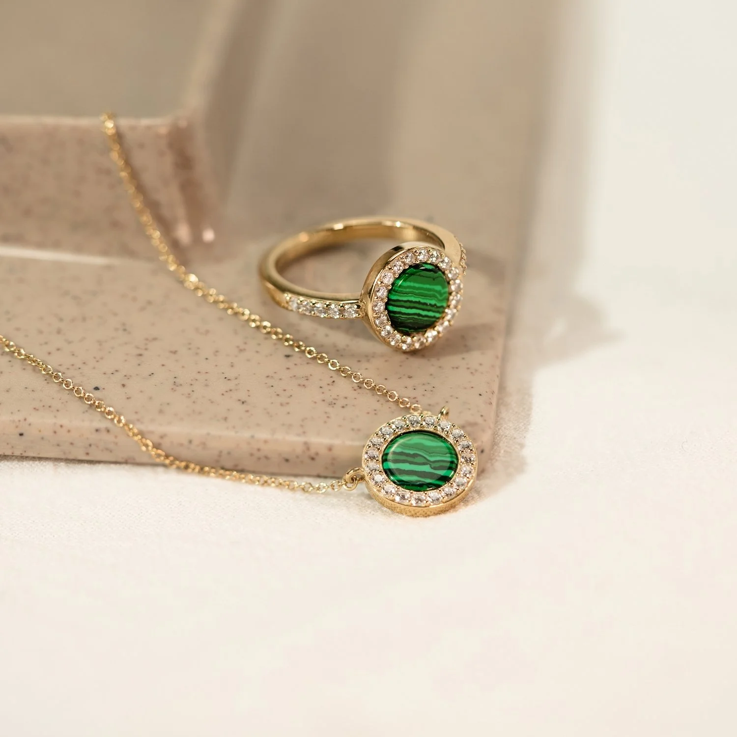 Gold jewelry set including a ring and necklace, both featuring green stone with black stripes and surrounded by small clear stones, placed on a light pink and white surface.