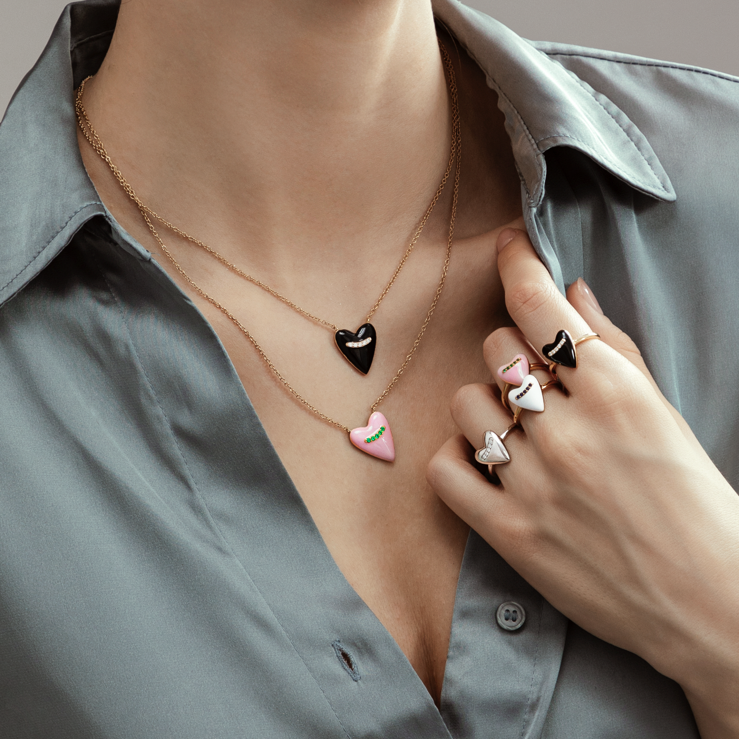 A woman wearing layered necklaces with heart-shaped pendants and multiple rings, dressed in a gray button-up shirt.