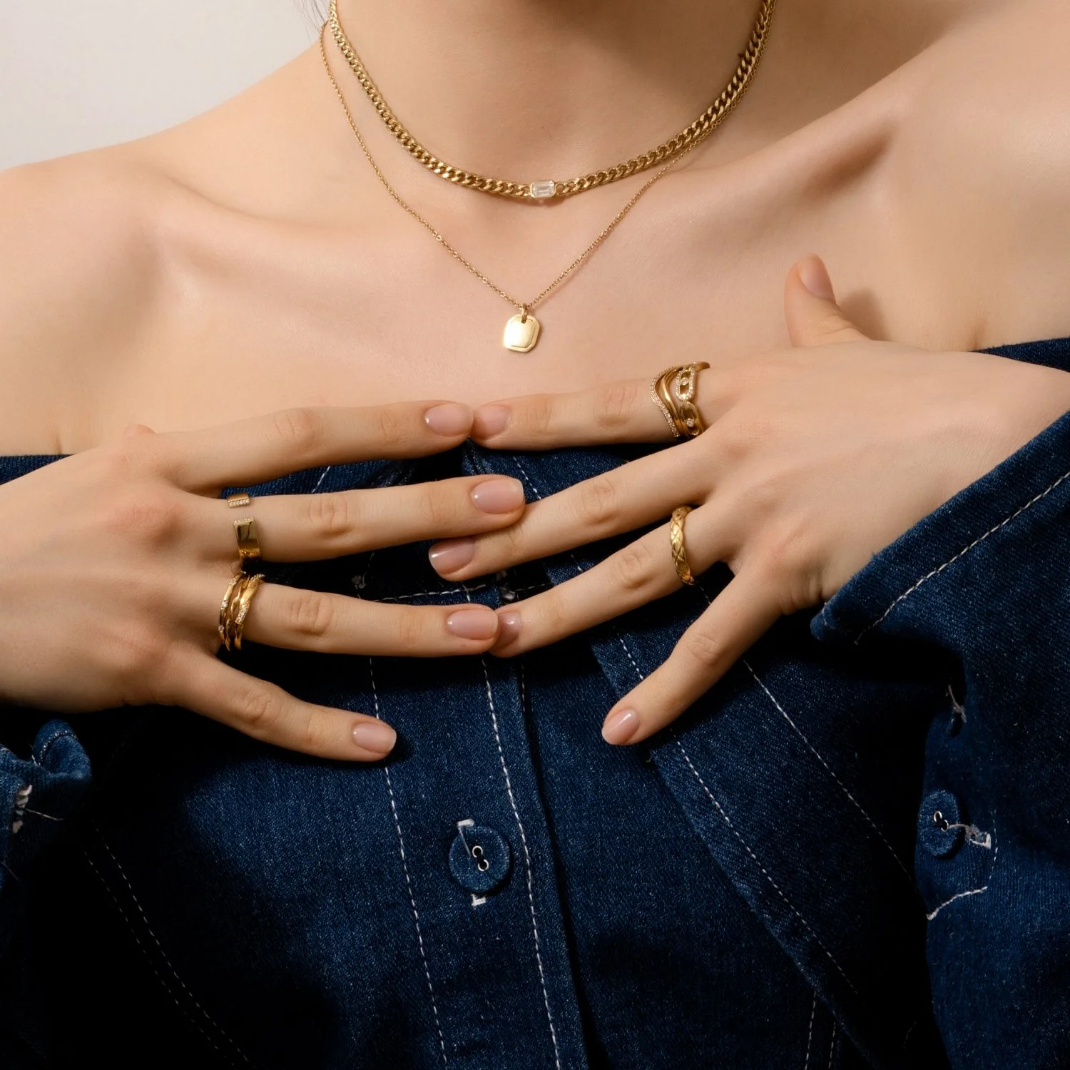 Close-up of a woman wearing multiple gold rings and layered gold necklaces, with her fingers resting on her chest and wearing a dark denim off-shoulder top.