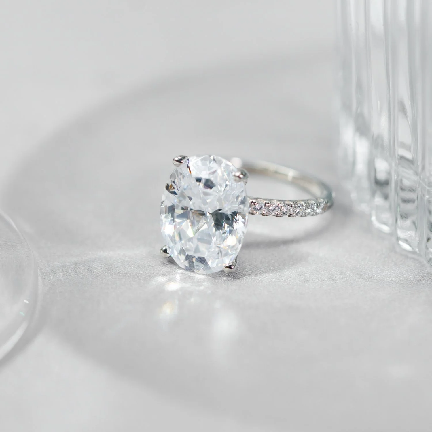 A silver ring with a large oval-shaped diamond and smaller diamonds on the band, placed on a white satin fabric surface next to a glass container and a clear glass block.