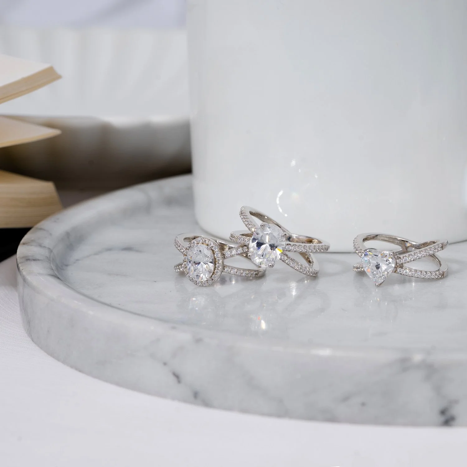 Three silver rings with large clear gemstones and smaller embedded stones on a round marble tray, with a white cup in the background.
