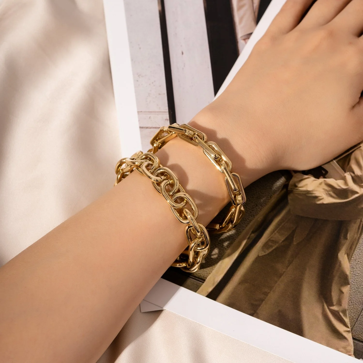 A person's wrist wearing two chunky gold chain bracelets resting on a white surface with a photograph underneath showing beige and black clothing.