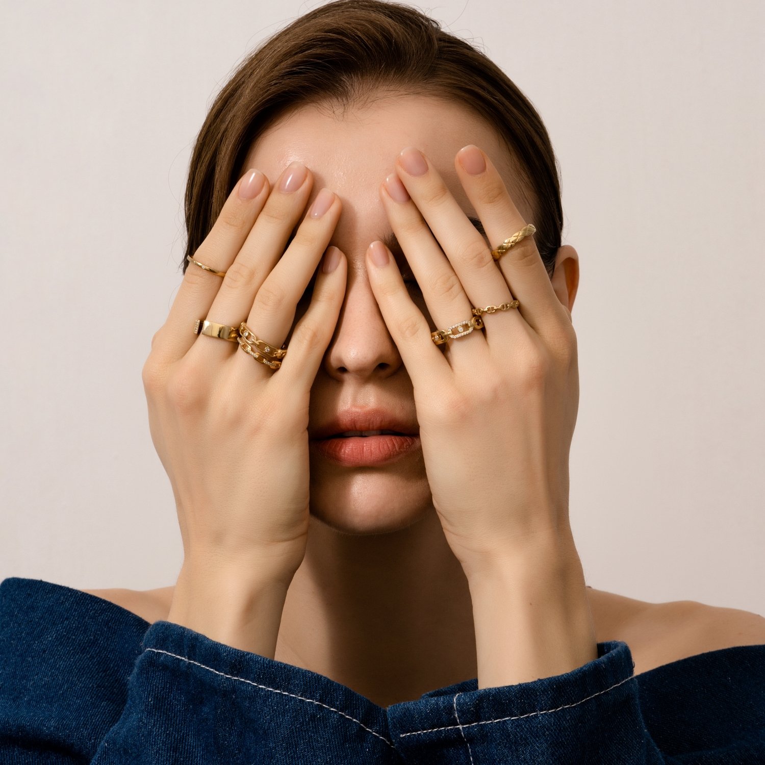 A woman with brown hair covering her face with both hands, wearing multiple gold rings on fingers, and a dark blue off-the-shoulder top.