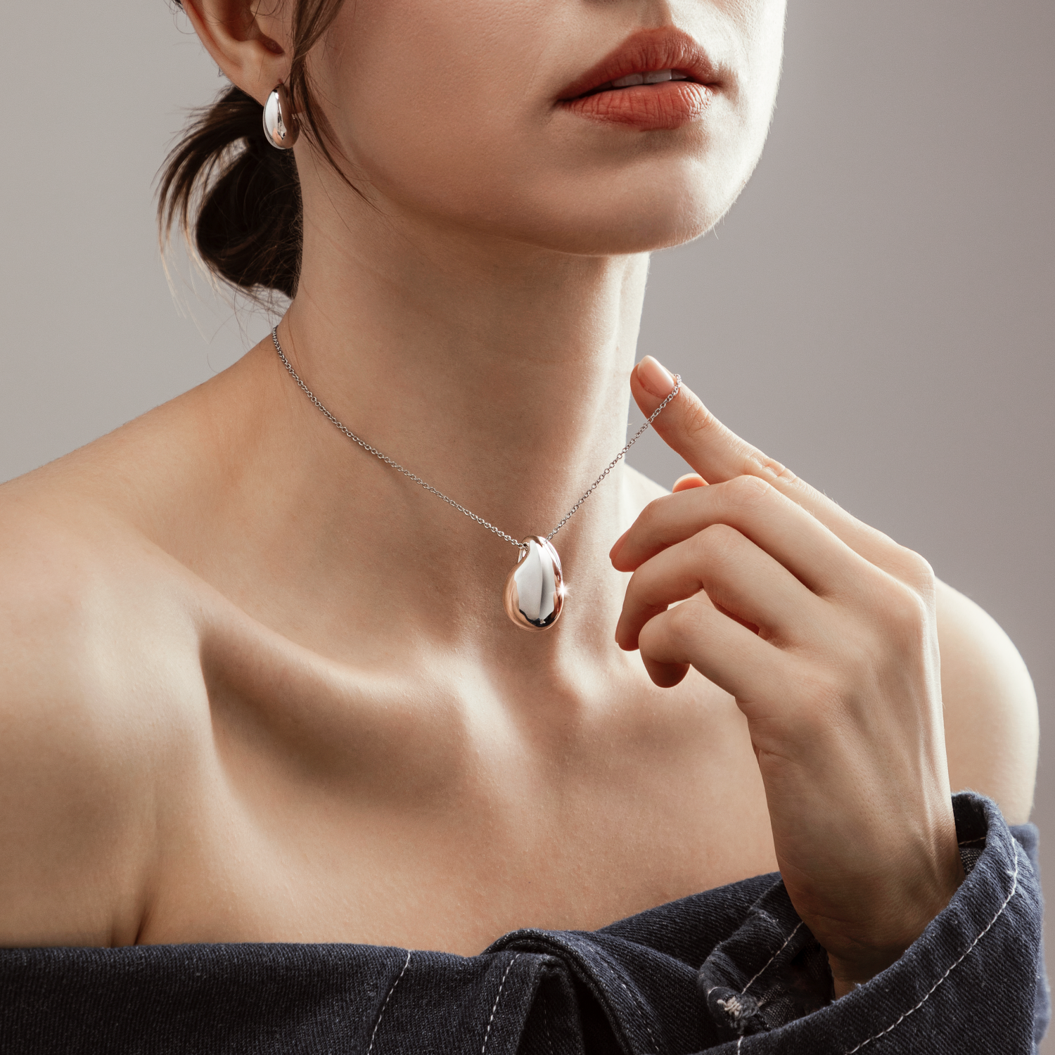 Close-up of a woman wearing silver jewelry, including a teardrop-shaped necklace and hoop earrings, with her hand gently touching the necklace.
