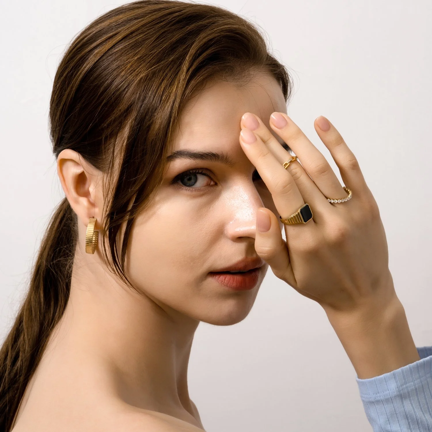 A young woman with brown hair and blue eyes, wearing gold earrings and rings, partially covering her face with her hand against a plain white background.