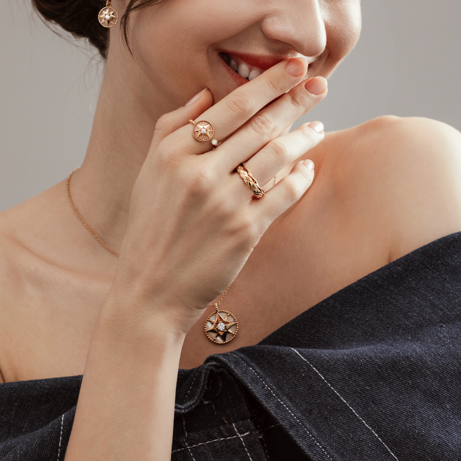 Woman wearing gold jewelry including earrings, necklace, and rings, with a black off-the-shoulder top, touching her lips with her fingers.